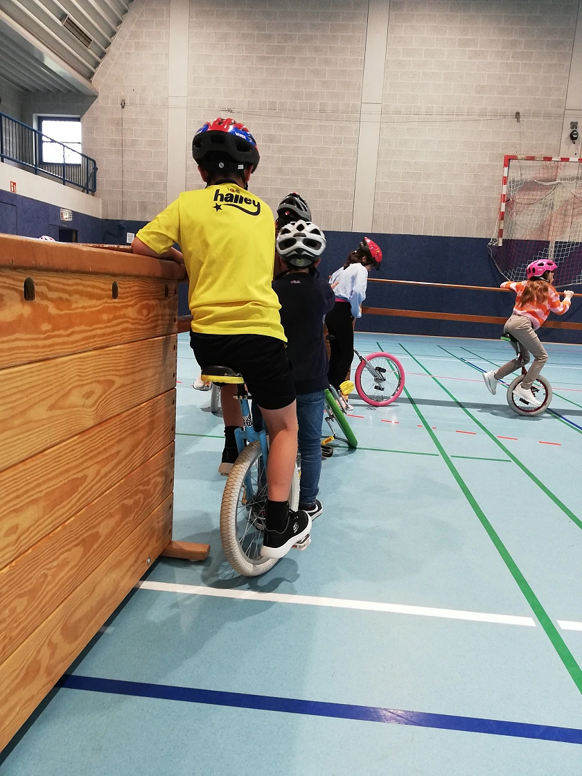 das Foto zeigt Kinder hintereinander auf einem Einrad. Alle Kinder tragen einen Fahrradhelm. Zum Festhalten dient ein großer Sprungkasten