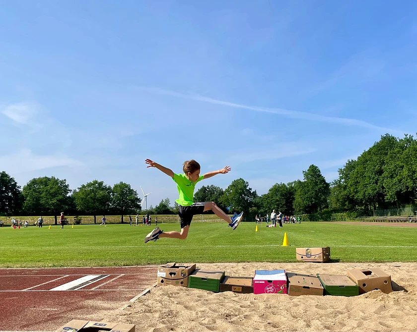 Das Foto zeigt ein Kind in der Luft, dass mit ausgestreckten Armen und Beinen über eine Weitsprunggrube springt